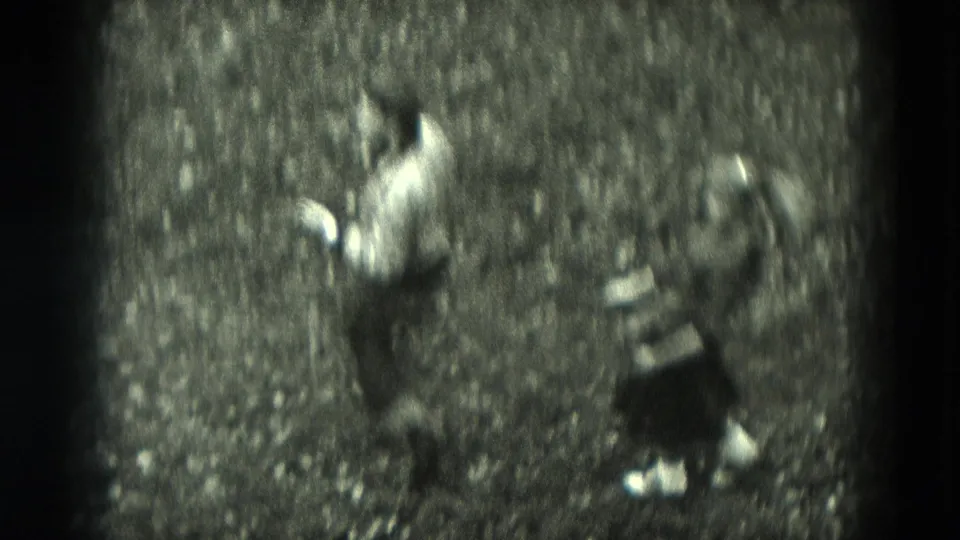 The couple appears mid-dance on an outdoor gravel path, with the man in a light shirt and the woman in a dark dress in black-and-white 8mm footage.
