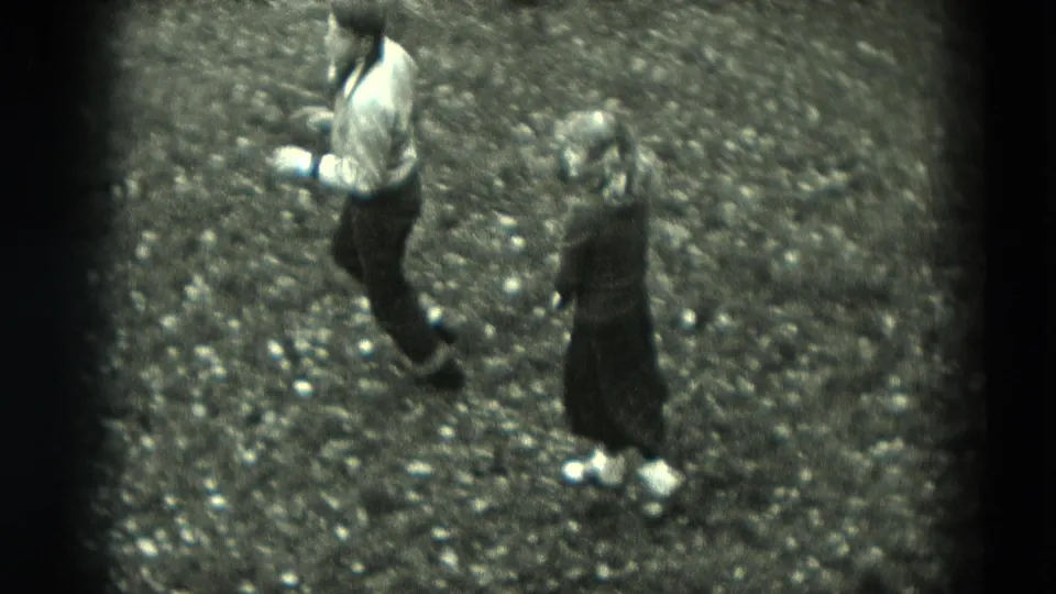 In the high-angle home-movie view, the man steps across the gravel while the woman in a dark dress stands nearby, suggesting a playful dance movement.