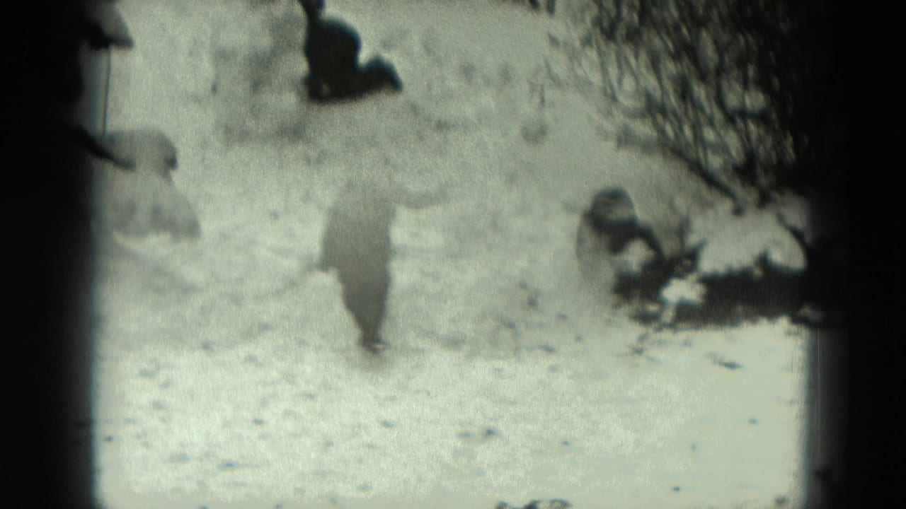 1951 Germany 8mm frame with a bundled figure moving across a snowy yard between small snowmen and winter shrubs.