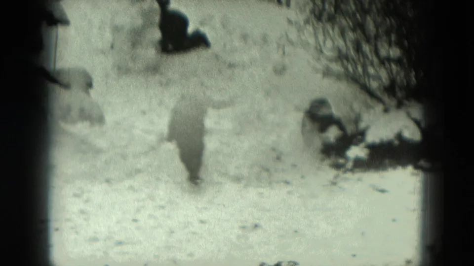 1951 Germany 8mm frame with a bundled figure moving across a snowy yard between small snowmen and winter shrubs.