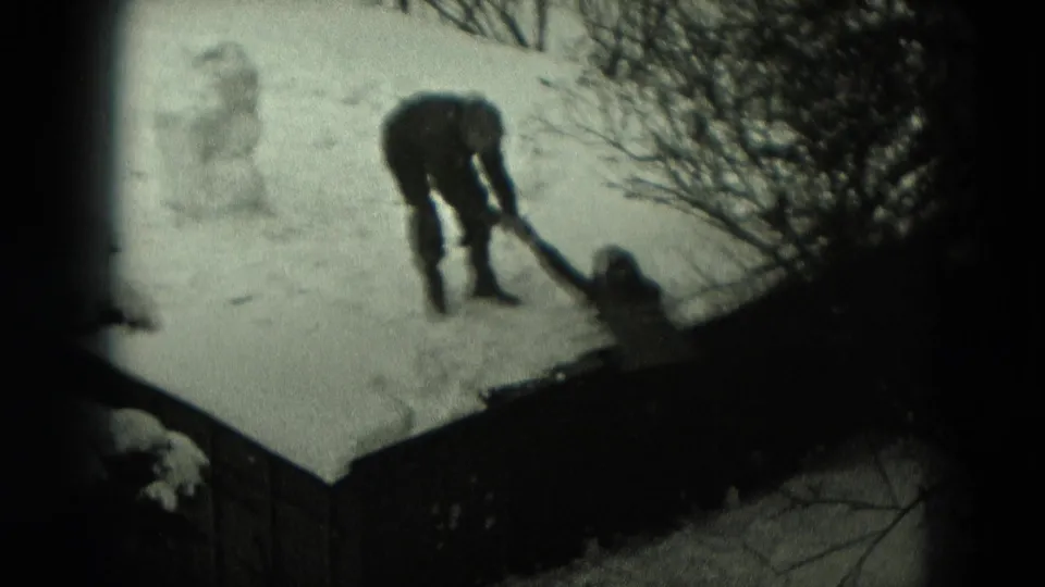 1951 Germany 8mm frame showing one bundled figure bending over snow near a fence, with another low figure partly visible nearby.