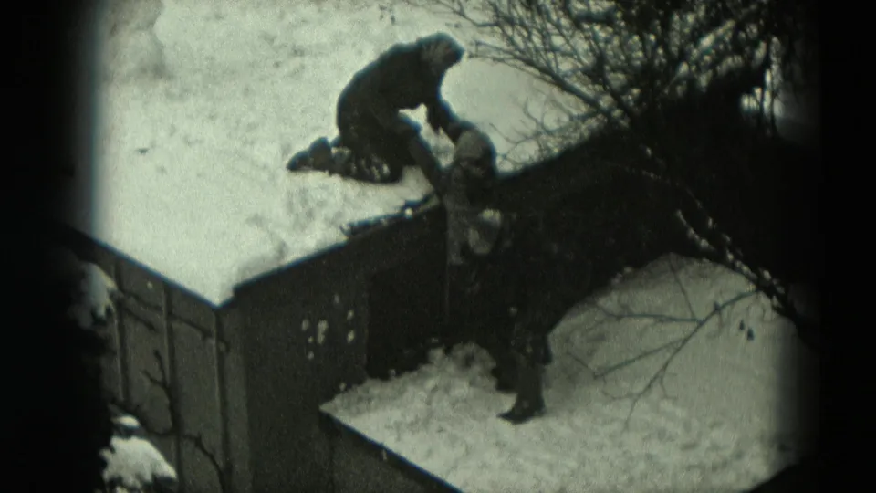 Home-movie frame from metadata-described 1951 Germany showing two bundled figures interacting across a snowy roof edge.