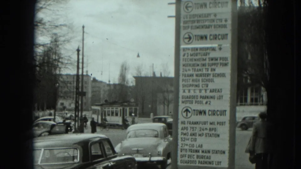 A West Germany street scene shows a directional town circuit sign, rounded cars, a trolley or tram, pedestrians, and overhead wires.