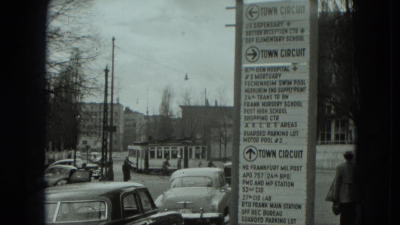 1951 West Germany street view with early cars, a tram in the distance, pedestrians, and a town-circuit sign listing local and U.S.-associated destinations.