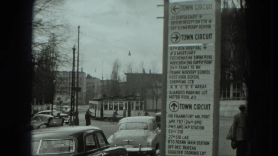 1951 West Germany street view with early cars, a tram in the distance, pedestrians, and a town-circuit sign listing local and U.S.-associated destinations.