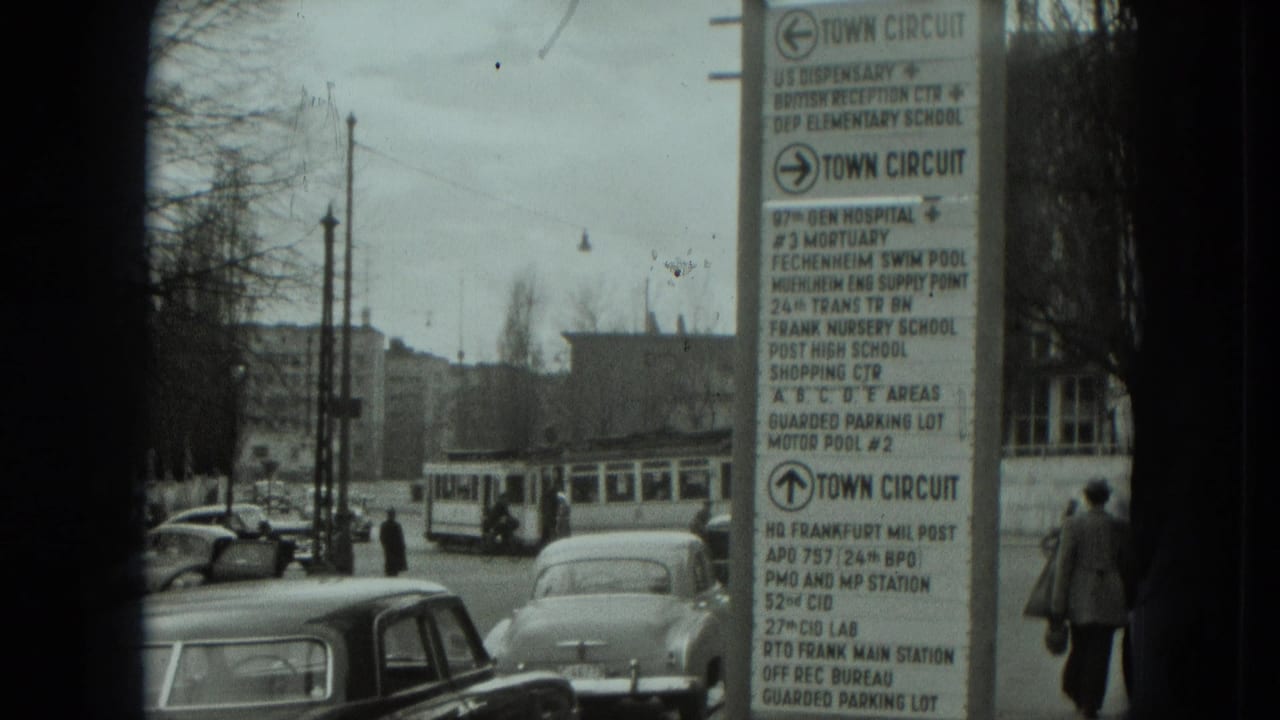 1951 West Germany street scene showing a tall town-circuit sign, parked classic cars, a tram, and pedestrians moving along the roadside.