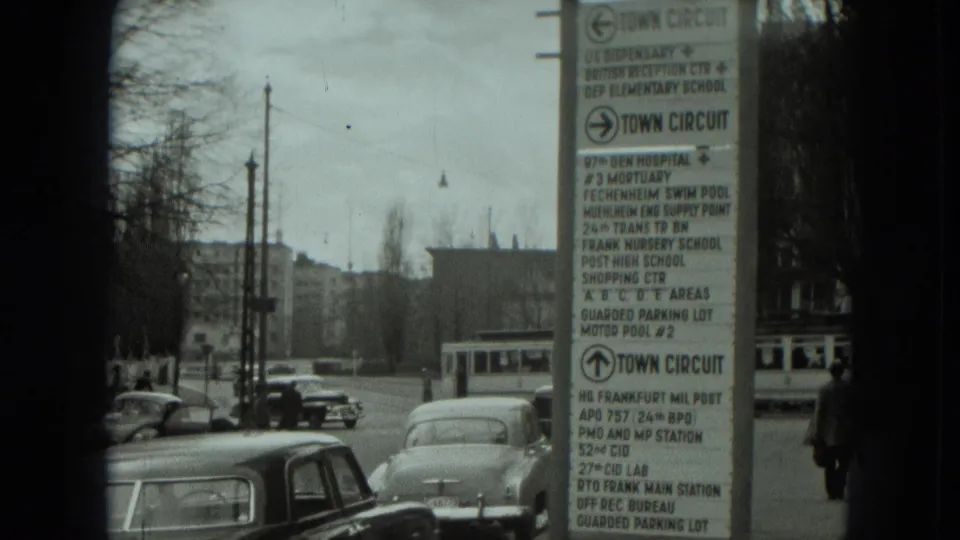 1951 West Germany street cutaway with parked cars, a tram partly visible, pedestrians, bare trees, and a tall sign naming local services and facilities.