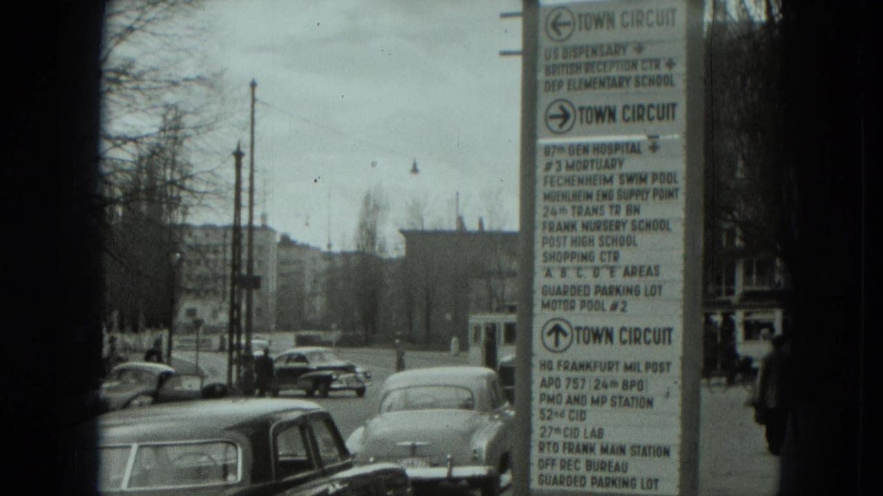 1951 West Germany street view from a moving vehicle, with a large directional sign, older cars, bare trees, and distant buildings.