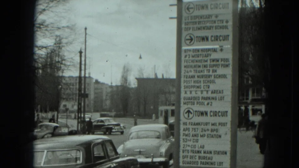 1951 West Germany street view from a moving vehicle, with a large directional sign, older cars, bare trees, and distant buildings.