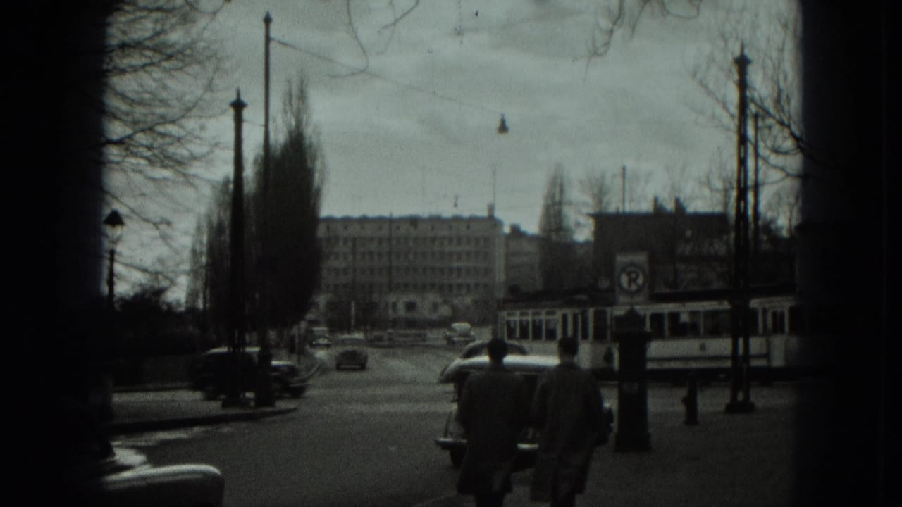 1951 West Germany intersection with pedestrians in coats, a trolley bus at right, cars on the road, overhead wires, and institutional buildings.