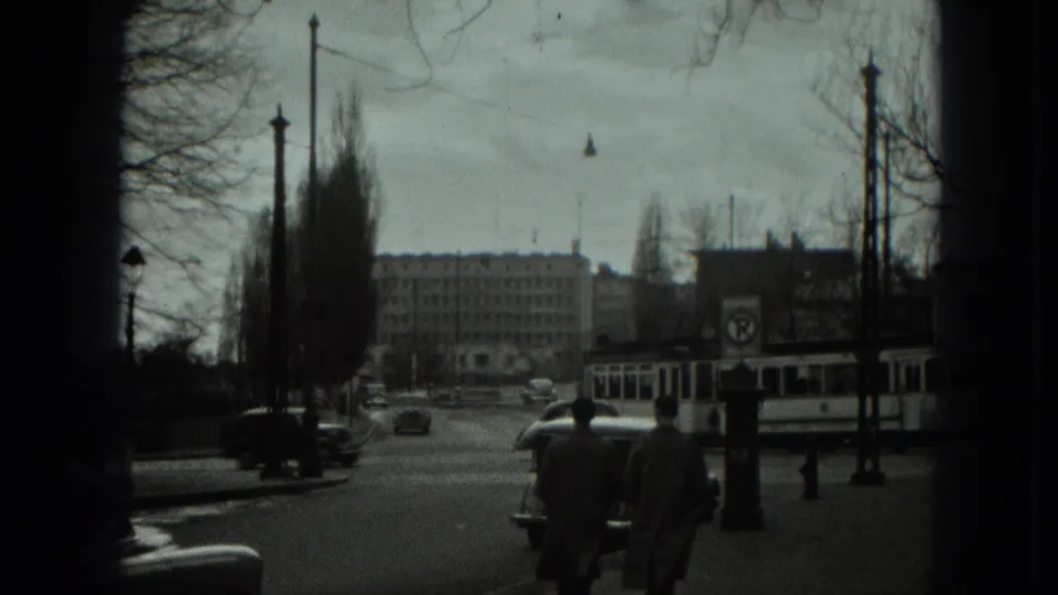 1951 West Germany intersection with pedestrians in coats, a trolley bus at right, cars on the road, overhead wires, and institutional buildings.