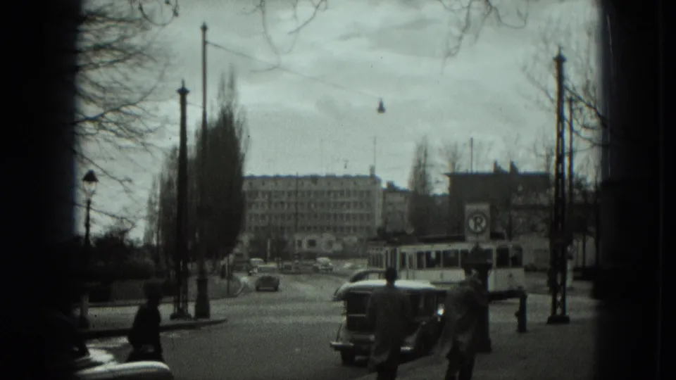 1951 West Germany street scene showing a trolley bus, pedestrians crossing or walking, cars, bare trees, and a large background building.