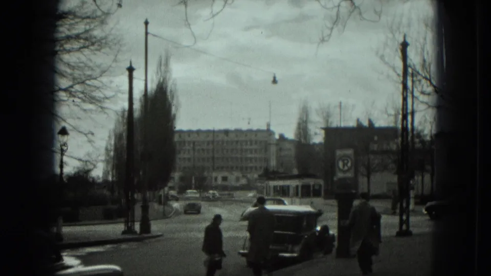 1951 West Germany intersection with pedestrians near the curb, a trolley bus, a dark automobile, bare trees, and overhead transit wires.