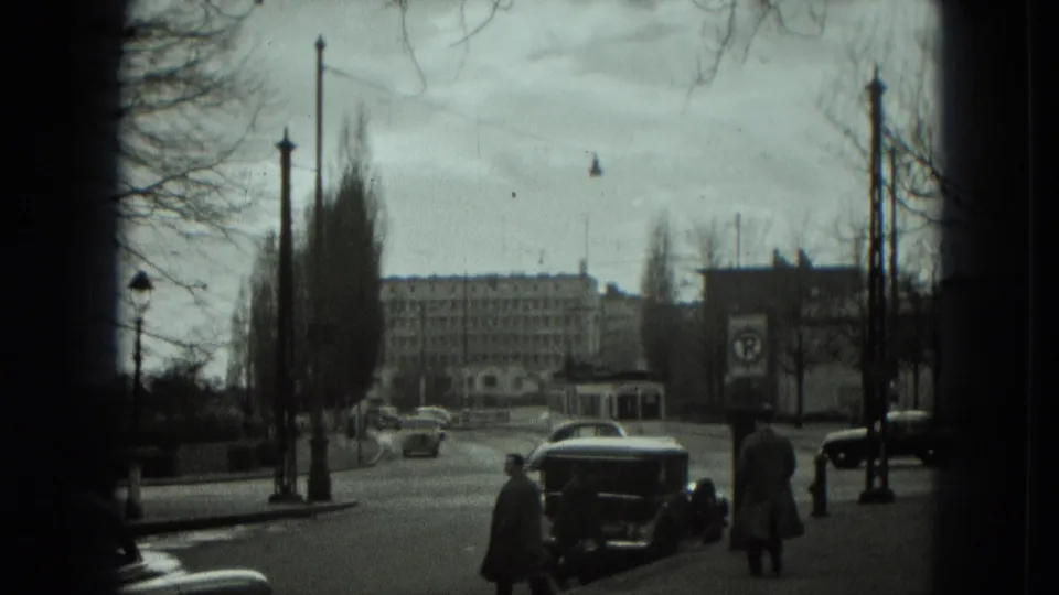 1951 West Germany street view with mid-century cars, pedestrians, bare trees, street lamps, and large buildings in the distance.