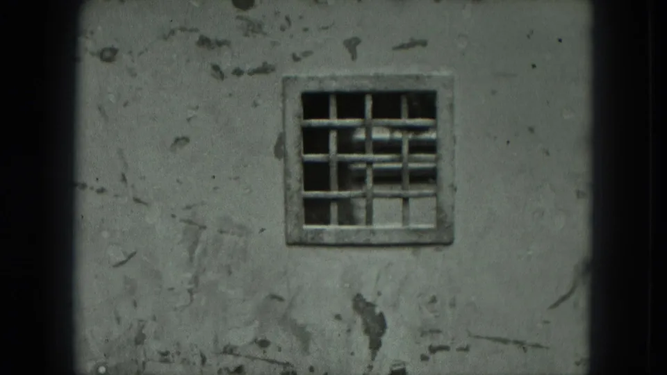 A centered barred window sits in a weathered cement wall, with chipped plaster and dark interior detail visible through the grid.