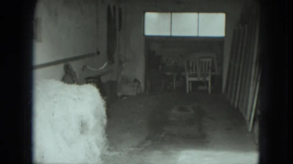 Shed-like private interior in 1951 Germany footage, showing hay, a chair, tools, boards, and bright window panes.