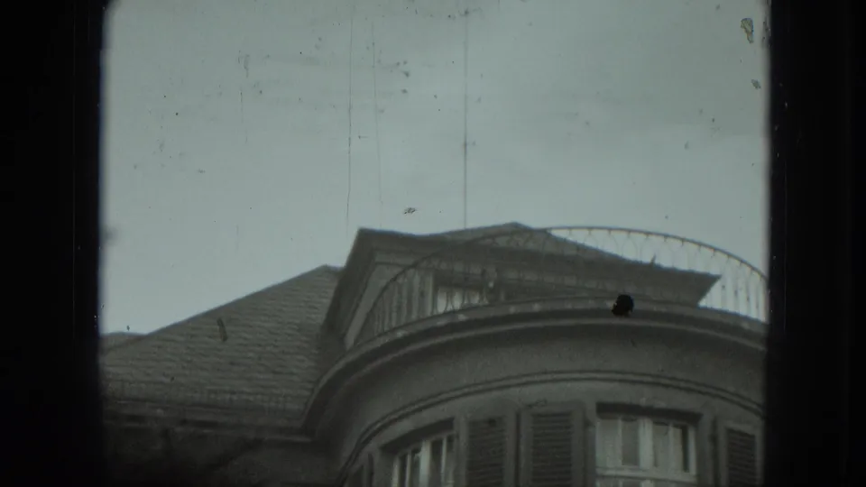 The camera frames a curved balcony, roof, shuttered windows, and cloudy sky in the 1951 West Germany exterior.