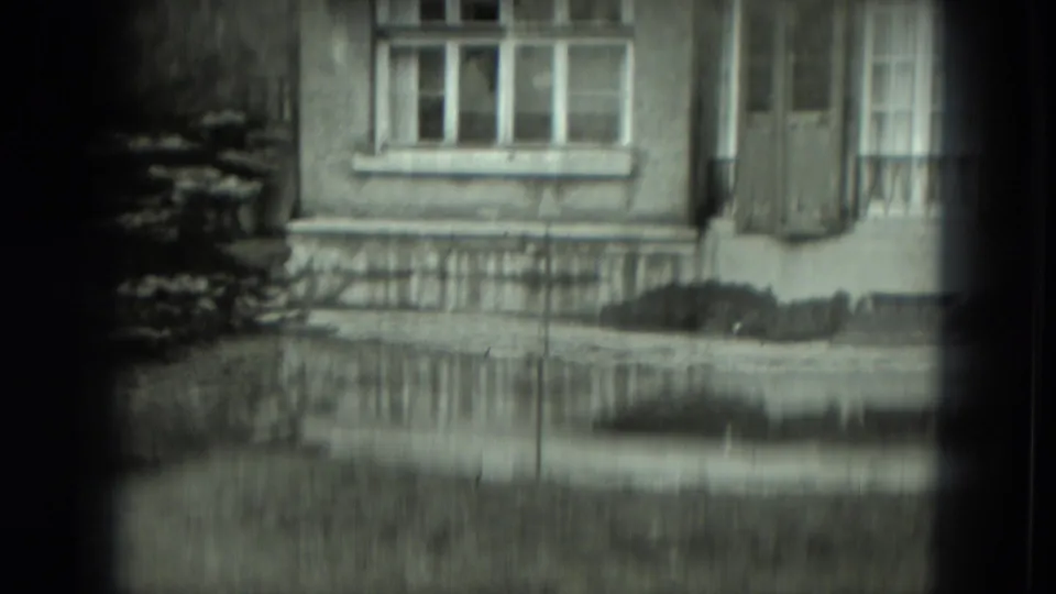 Soft black-and-white 1951 Germany 8mm frame of a house facade with windows, shutters, railing, and lawn.