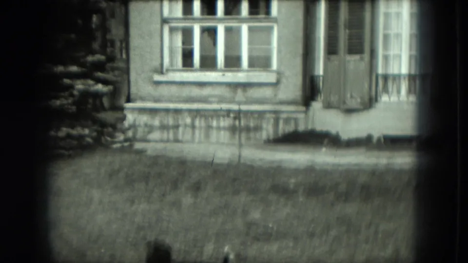 Black-and-white 8mm view of a German house exterior with shuttered windows, railing, and grassy foreground.