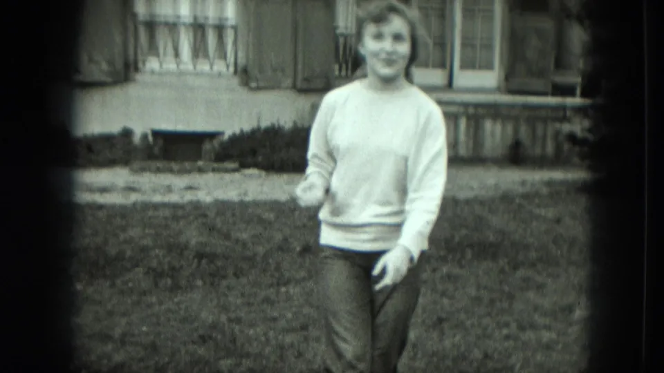 A smiling woman in a light sweater and dark pants stands in a grassy residential yard in Germany, with a house facade behind her.