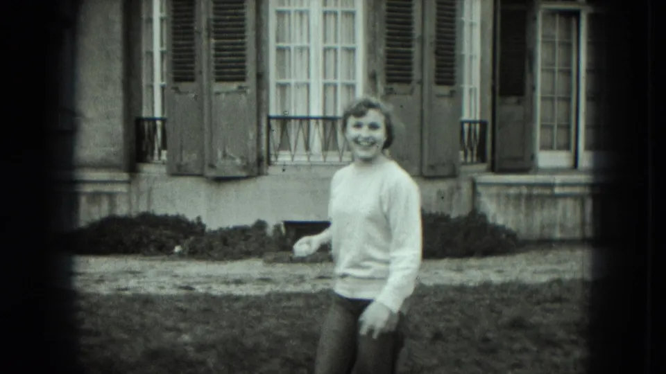 A woman smiles toward the camera in a grassy yard, with shuttered windows and residential architecture visible behind her.