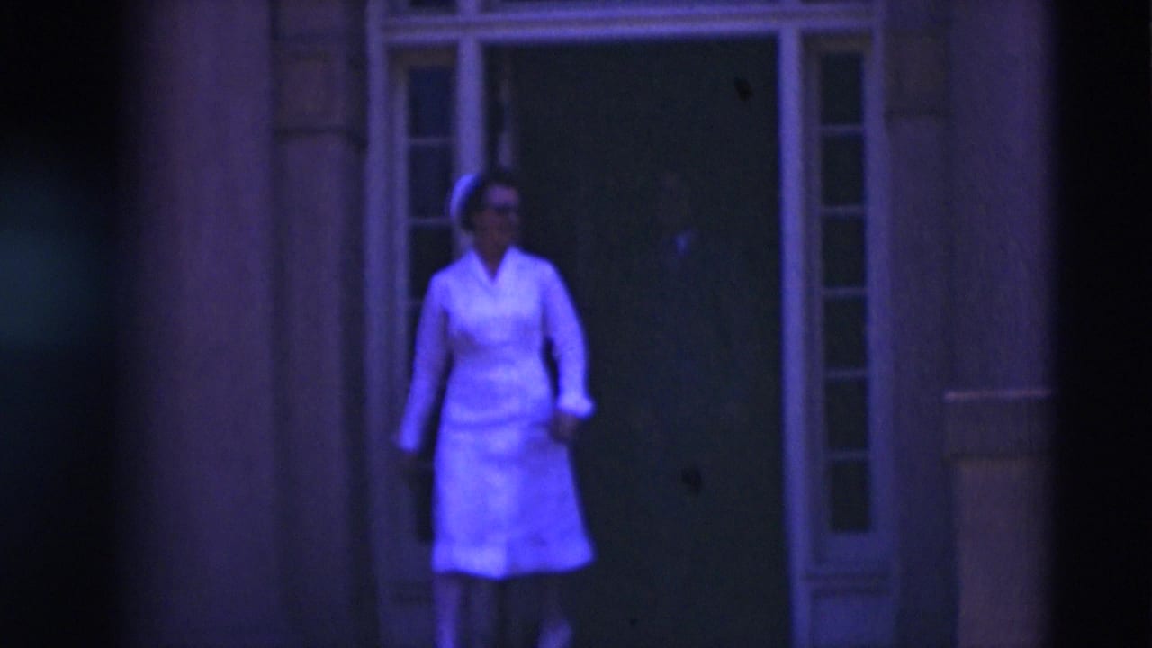 Woman in a white nurse-style uniform walks out of a shaded doorway.
