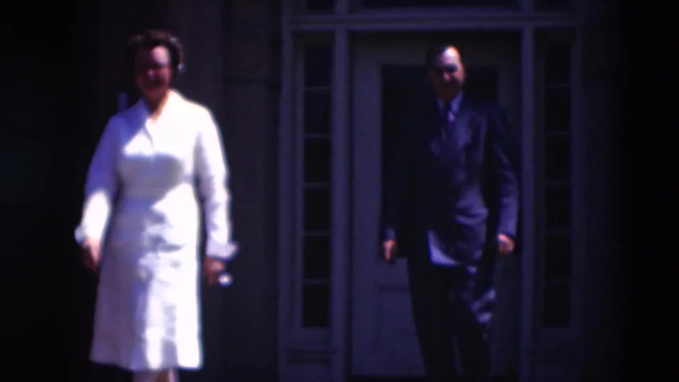 The 1951 8mm frame shows the woman in white walking toward camera as the man in a dark suit follows from the entrance.