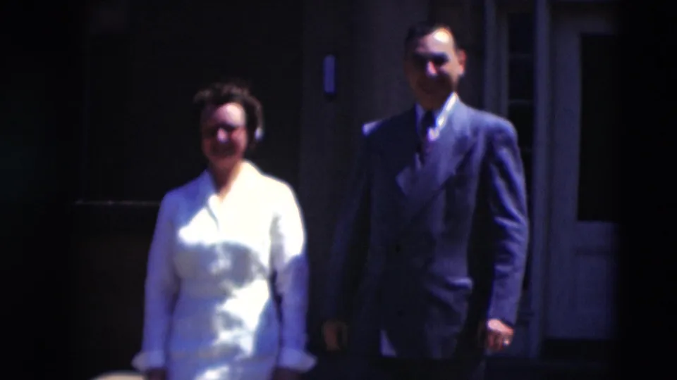 A closer 1951 home-movie frame shows the woman in white and the suited man walking toward the camera outside the building.