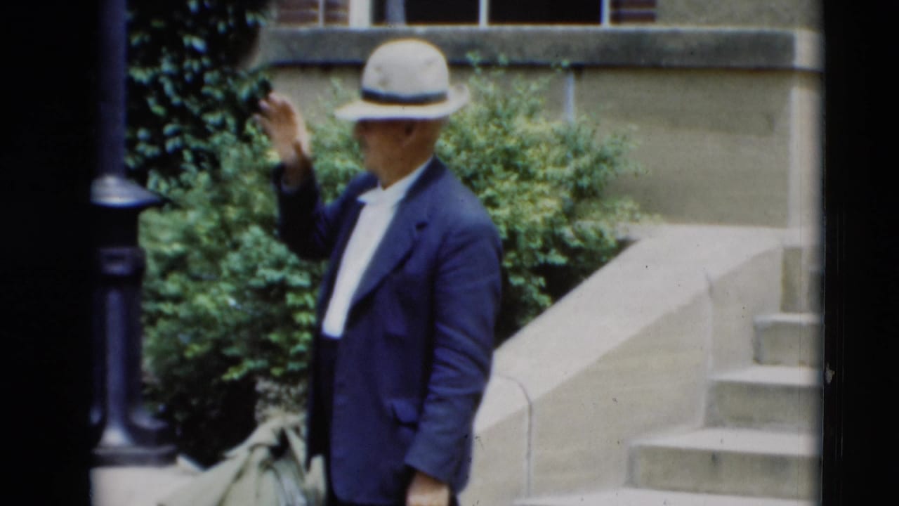 Man in fedora raises one hand while standing near steps and shrubs.