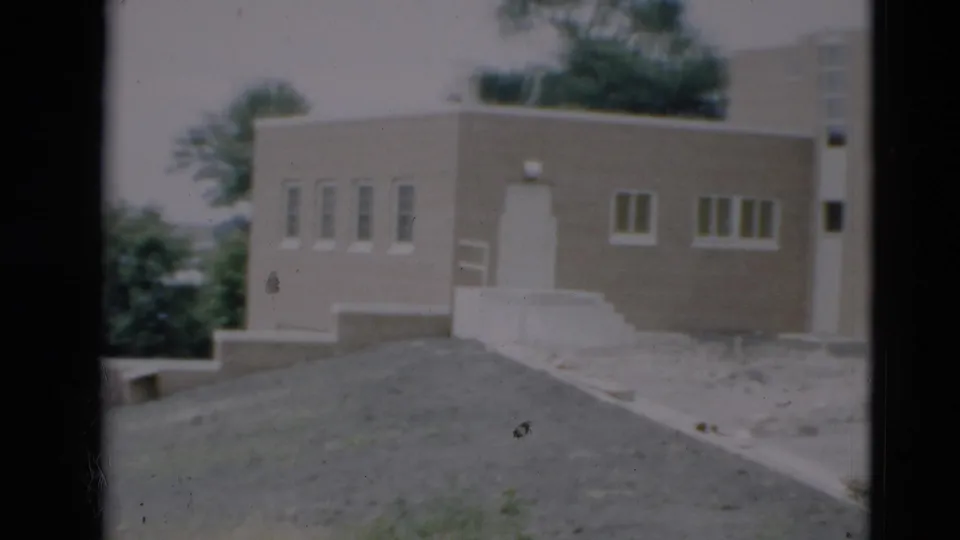 1951 Paducah home-movie frame showing a pale flat-roof building, white-trimmed windows, trees, and a gravel hillside.