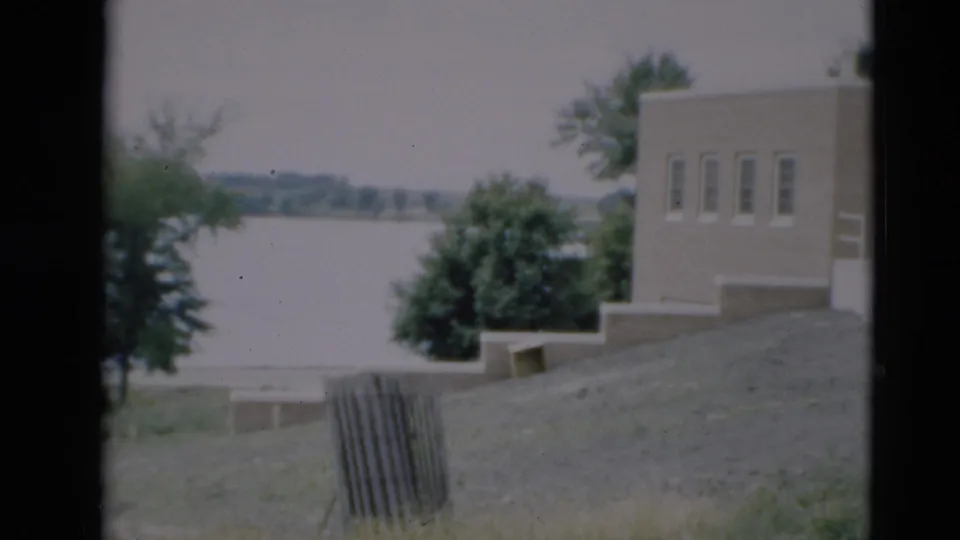 1951 Paducah home-movie frame showing a sloped riverside area, trees, water, and the edge of a flat-roof building.
