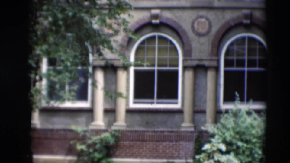 1951 8mm view of a Paducah facade with arched windows, columns, a brick base, branches, shrubs, and soft film grain.