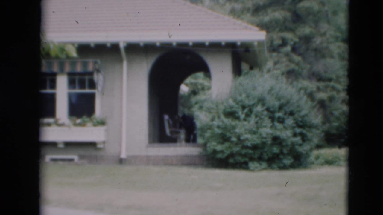 1951 Paducah home-movie frame showing a grey residential exterior with arched porch, divided window, lawn, and shrubs.