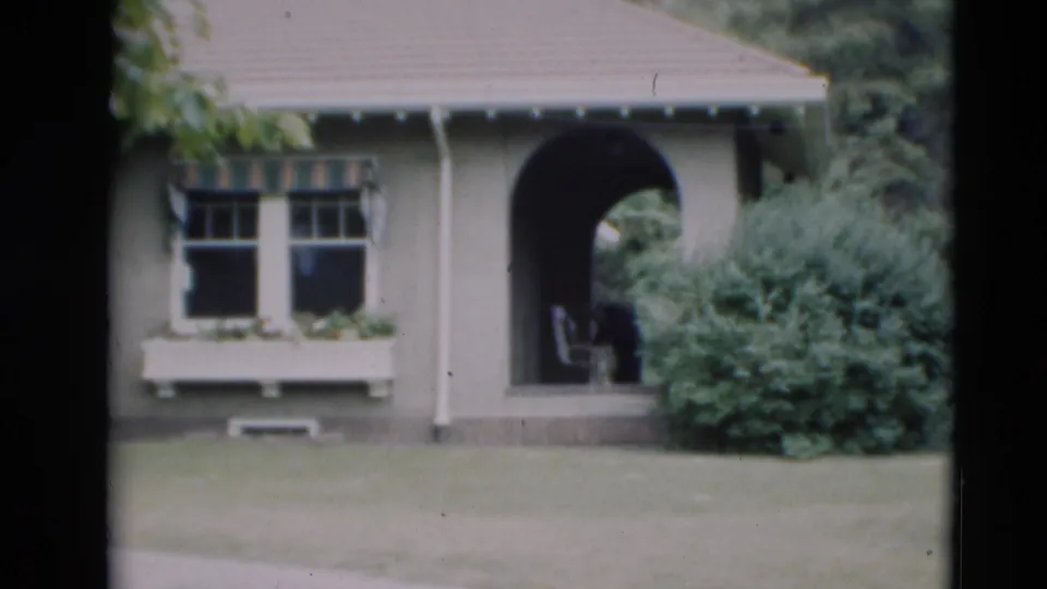 1951 Paducah exterior view of a grey house with an arched entry, window box, shrubs, and an indistinct figure in the porch shade.