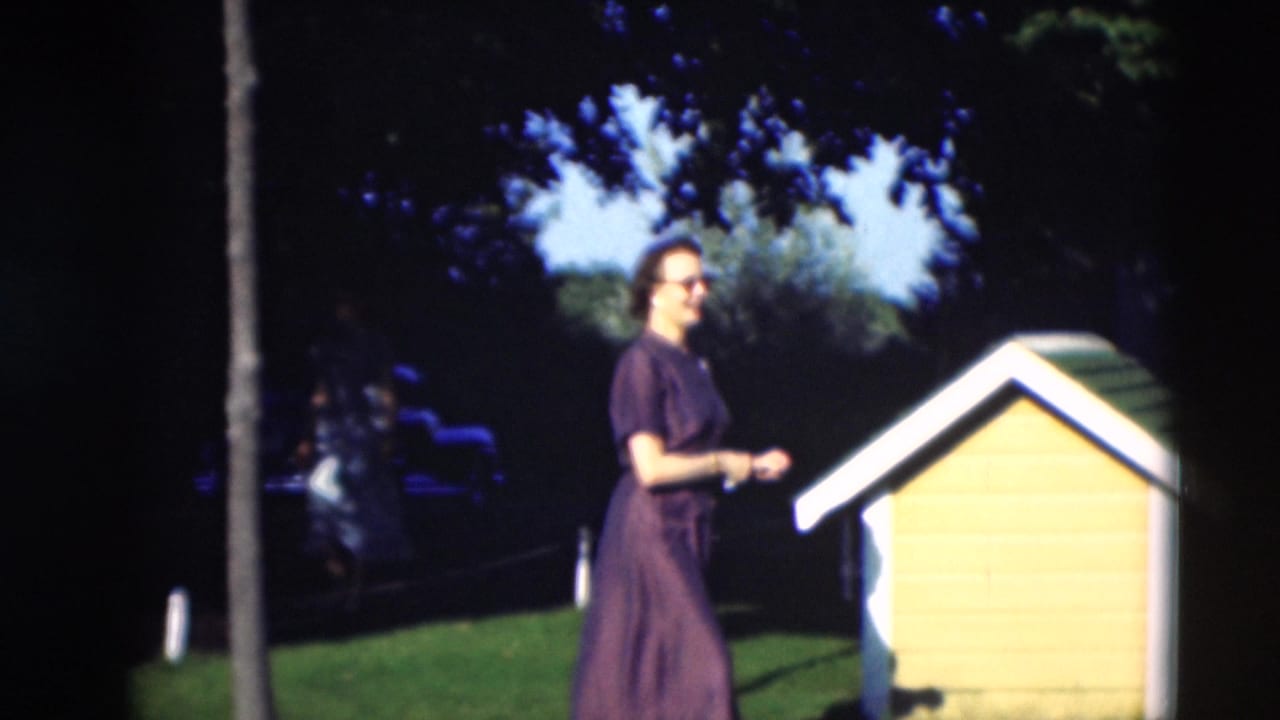 A woman in a dark dress stands beside a yellow and white dog house in a sunny yard in the 1951 Paducah home movie.