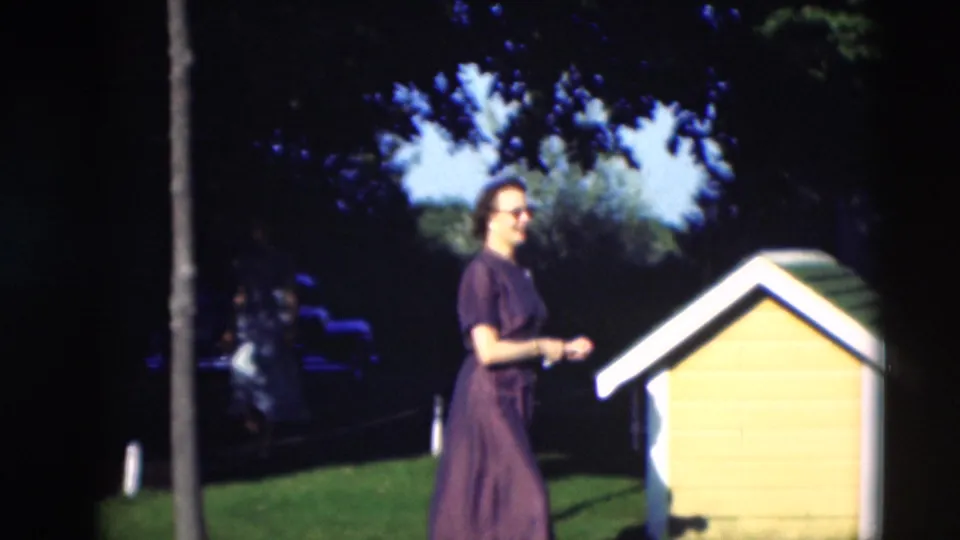 A woman in a dark dress stands beside a yellow and white dog house in a sunny yard in the 1951 Paducah home movie.