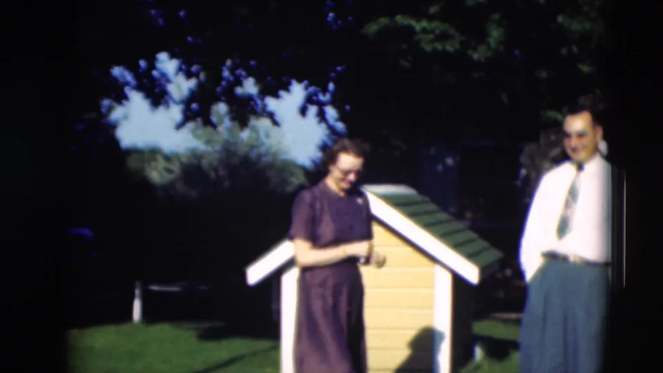 The woman looks down near the dog house while an older man in a white shirt and tie stands beside her in the yard.
