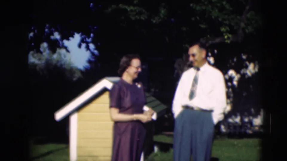 An older couple stands together beside the yellow and white dog house in the Paducah yard, with trees and shade behind them.