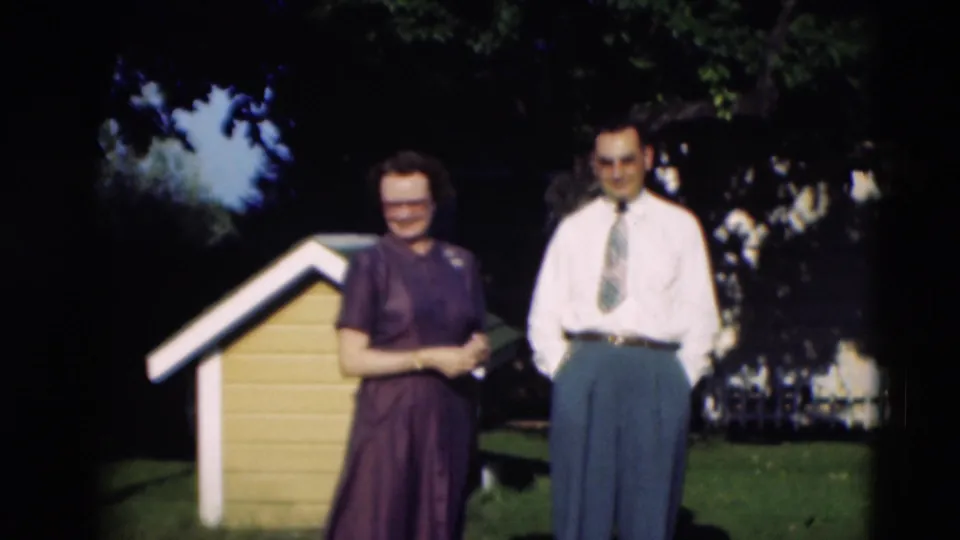 1951 Paducah home-movie frame showing an adult couple standing near a yellow doghouse under trees in a backyard.