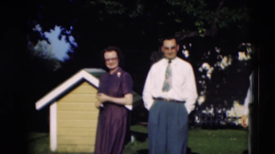 1951 Paducah home-movie still of a smiling woman and man posed near a small yellow doghouse in daylight.