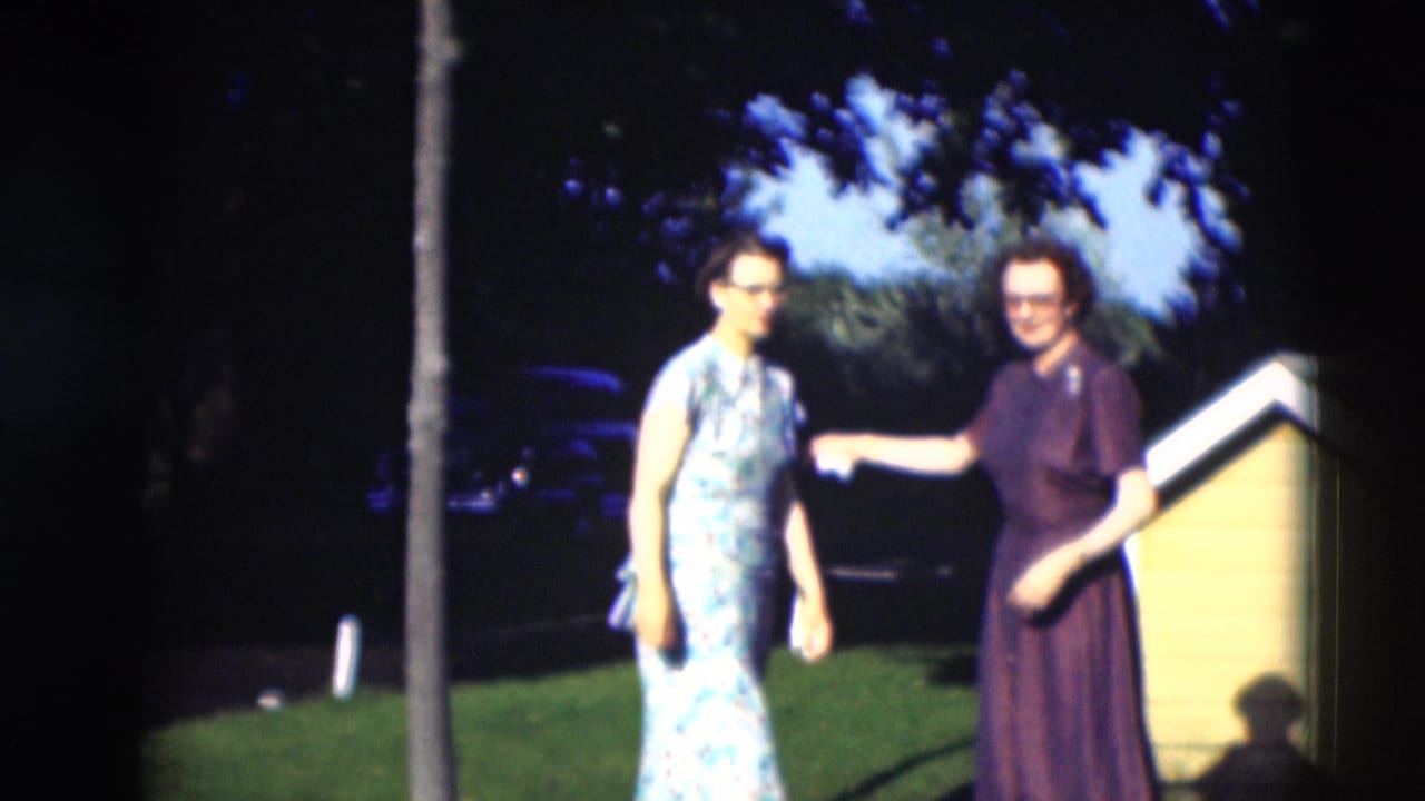 Two women stand and gesture on a Paducah lawn, with a yellow house edge and shaded trees behind them.