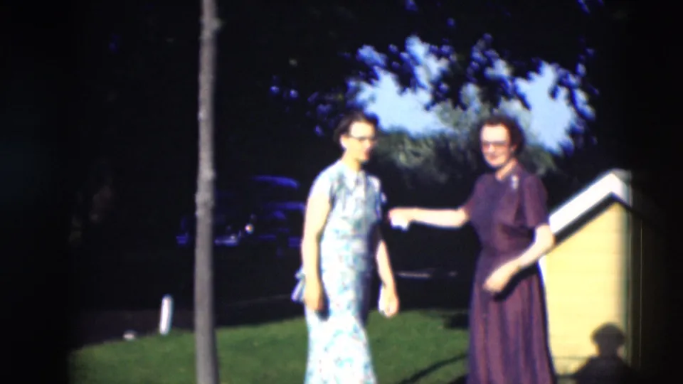 Two women stand and gesture on a Paducah lawn, with a yellow house edge and shaded trees behind them.