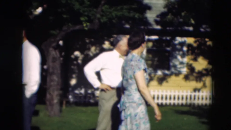 Adults in mid-century clothing stand and move across a residential lawn with a white picket fence in the background.