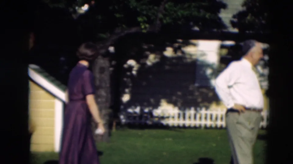 In 1951 Paducah home-movie footage, a woman in a purple dress and a man in a white shirt stand on a backyard lawn near a fence and trees.