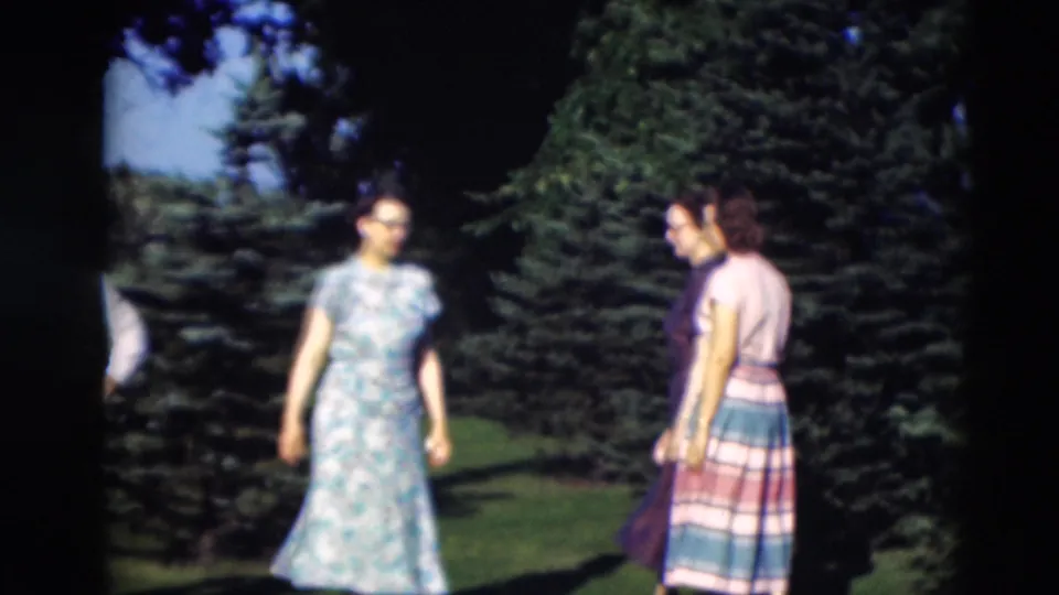 A woman in a floral dress approaches two women near evergreen trees during a 1951 Paducah backyard gathering.