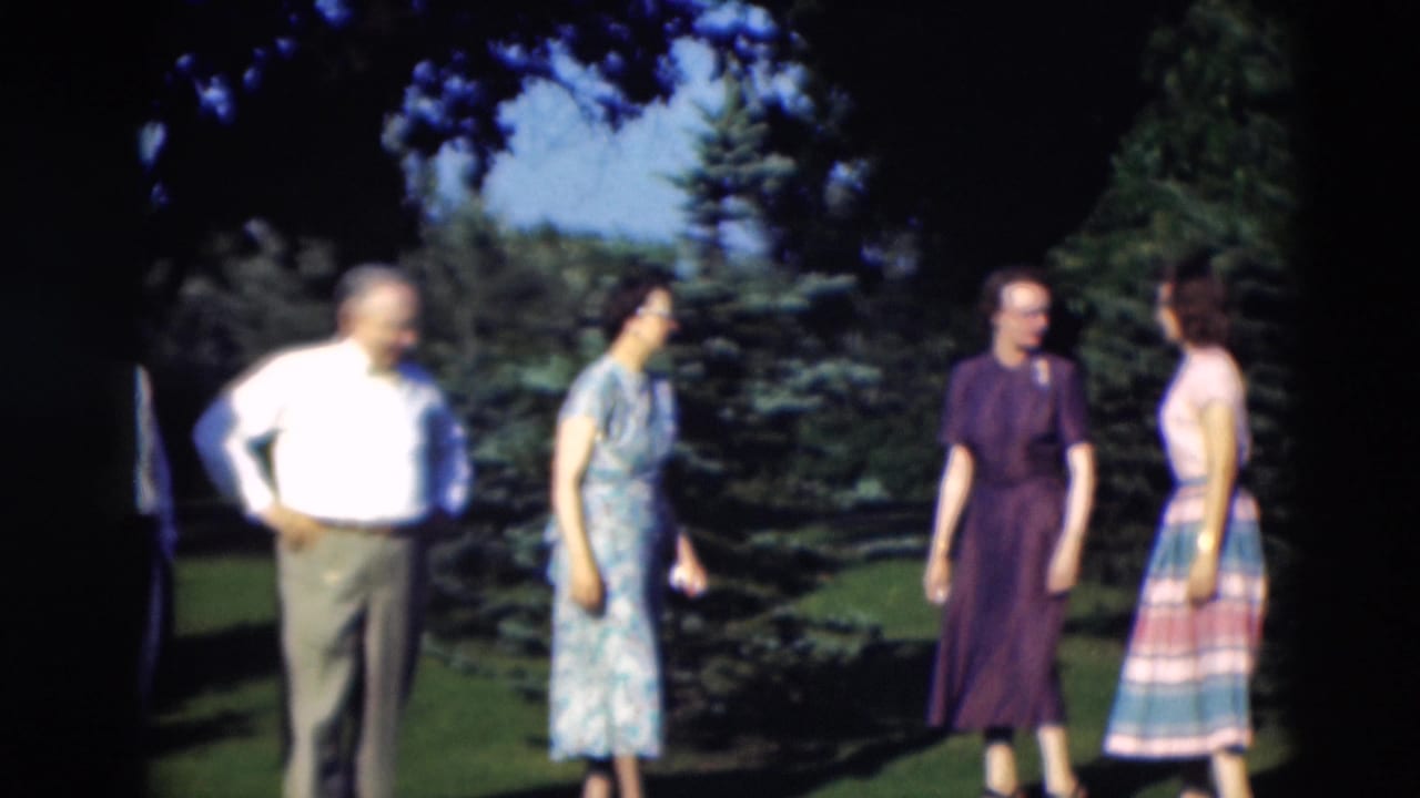 Adults stand on a sunny lawn near trees, with men in shirts and women in dresses.