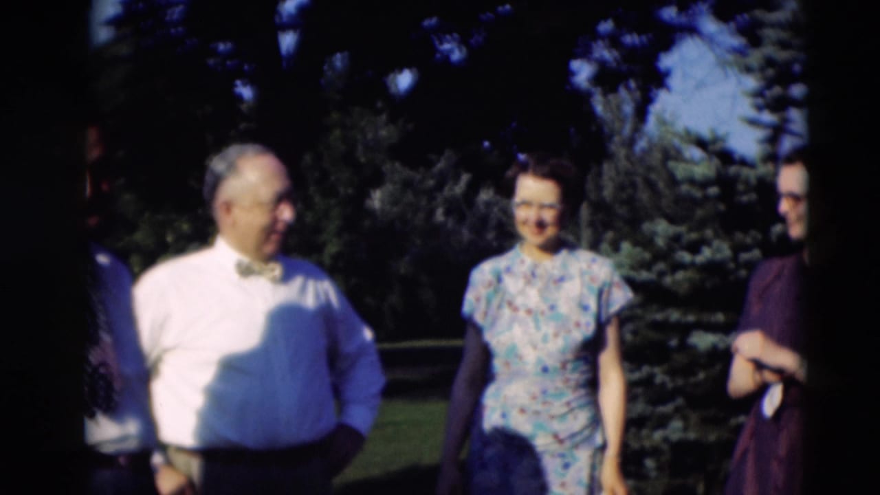 In the supplied 1951 Paducah home-movie frame, adults with glasses stand in bright sun beside evergreen trees on a lawn.
