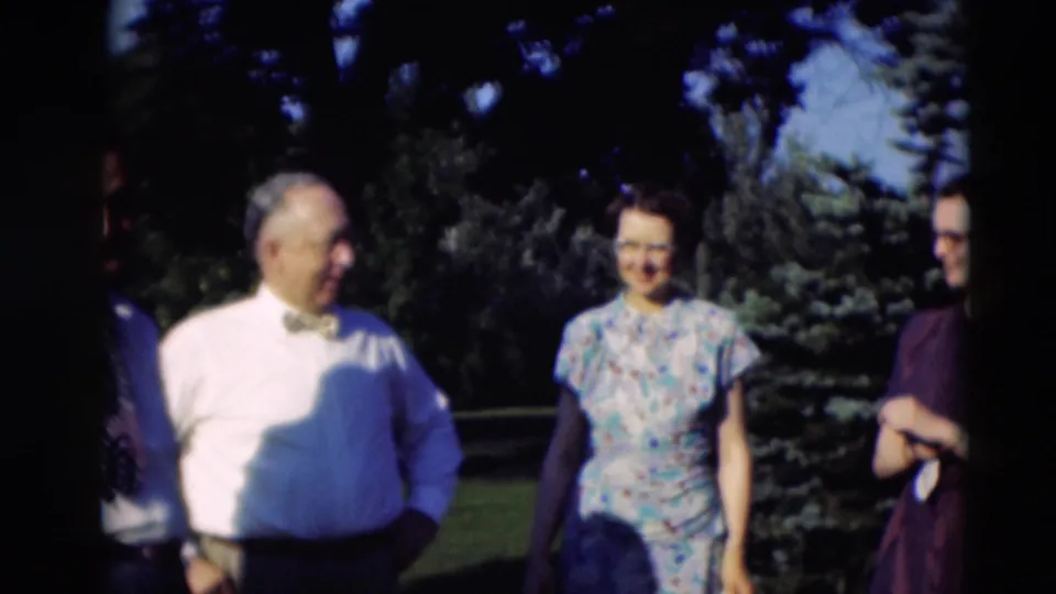 In the supplied 1951 Paducah home-movie frame, adults with glasses stand in bright sun beside evergreen trees on a lawn.