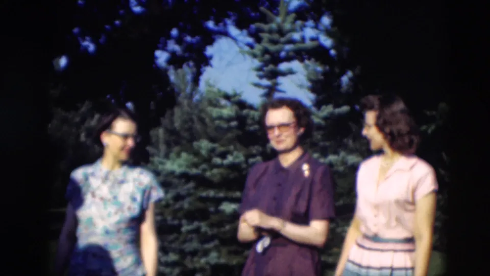 Three adults in the supplied 1951 Paducah home movie stand together on a lawn, smiling and conversing beside evergreen trees.