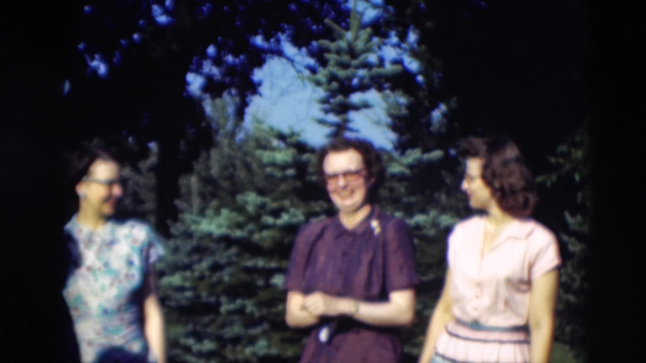 Three women in dresses stand outdoors near evergreen trees, two smiling toward the camera.